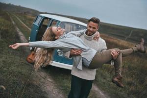 leven is geweldig knap jong Mens draag- zijn aantrekkelijk vriendin en glimlachen terwijl staand in de buurt de blauw retro stijl mini busje foto