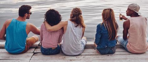 aan het doen wat dan ook ze willen. achterzijde visie van jong mensen in gewoontjes slijtage pratend terwijl zittend Aan de pier foto