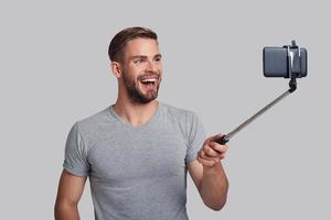 handig manier naar nemen selfie. knap jong glimlachen Mens nemen selfie terwijl staand tegen grijs achtergrond foto