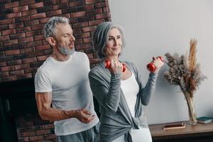 modern senior paar in sport- kleding oefenen en gebruik makend van hand- gewicht terwijl uitgeven tijd Bij huis foto