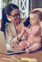 zeggen Hallo naar vader weinig baby meisje pratend Aan mobiel telefoon terwijl zittend Aan kantoor bureau met haar moeder in kantoor foto