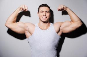 tonen zijn perfect spieren. jong gespierd Mens tonen zijn biceps en glimlachen terwijl staand tegen grijs achtergrond foto