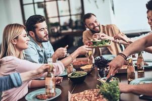 hebben een Super goed tijd. groep van jong mensen in gewoontjes kleding aan het eten en glimlachen terwijl hebben een avondeten partij binnenshuis foto