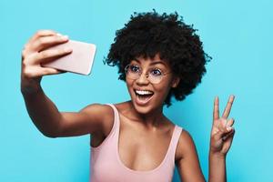 sharing mooi hoor foto's. aantrekkelijk jong Afrikaanse vrouw glimlachen en nemen selfie terwijl staand tegen blauw achtergrond foto