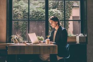 vol concentratie Bij werk. zelfverzekerd jong Mens in slim gewoontjes slijtage op zoek Bij laptop terwijl zittend in de buurt venster in creatief kantoor of cafe foto