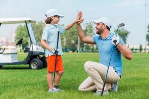 mijn weinig winnaar vrolijk jong Mens en zijn zoon geven high-five naar elk andere terwijl staand Aan de golf Cursus foto