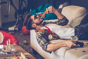 nog steeds kreeg energie naar feest. jong knap Mens in zonnebril aan het eten pizza en Holding bedieningshendel in zijn hand- terwijl aan het liegen Aan sofa in rommelig kamer na partij foto