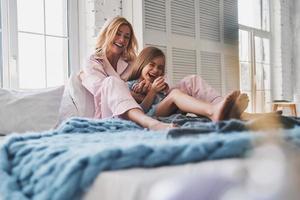 genieten van mooi hoor ochtend- samen. mooi jong moeder omarmen haar dochter en glimlachen terwijl zittend Aan de bed Bij huis foto