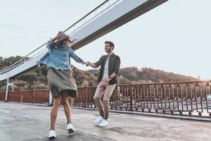 liefde is in de lucht. vol lengte van speels jong paar Holding handen en spinnen terwijl dansen Aan de brug buitenshuis foto