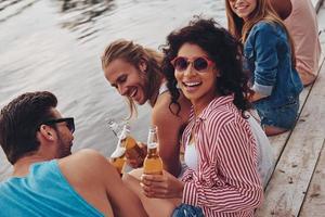zomer plezier. groep van gelukkig jong mensen in gewoontjes slijtage glimlachen en drinken beer terwijl zittend Aan de pier foto