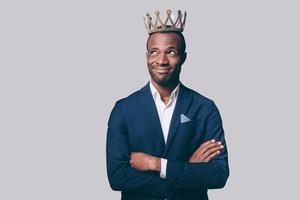 koning knap jong Afrikaanse Mens in kroon en slim gewoontjes jasje maken een gezicht terwijl staand tegen grijs achtergrond foto