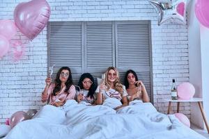 zo veel pret vier aantrekkelijk jong Dames in pyjama drinken cocktails terwijl aan het liegen in de bed met ballonnen allemaal over- de kamer foto