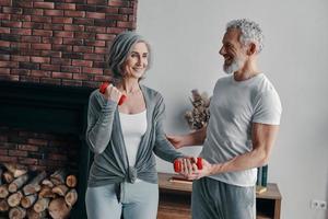 vrolijk senior paar in sport- kleding oefenen en gebruik makend van hand- gewicht terwijl uitgeven tijd Bij huis foto