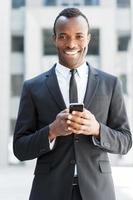 zakenman met mobiel telefoon. vrolijk jong Afrikaanse Mens in formele kleding Holding mobiel telefoon en glimlachen terwijl staand buitenshuis foto