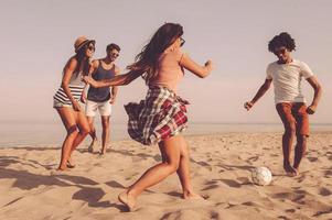 het is de mijne groep van vrolijk jong mensen spelen met voetbal bal Aan de strand met zee in de achtergrond foto