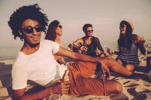 genieten van strand partij met vrienden. vrolijk jong mensen uitgeven mooi hoor tijd samen terwijl zittend Aan de strand en drinken bier foto