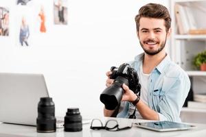 zelfverzekerd fotograaf. vrolijk jong Mens Holding digitaal camera en glimlachen terwijl zittend Bij zijn werken plaats foto