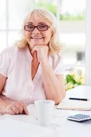 krijgen financiën Rechtdoor. gelukkig senior vrouw Holding hand- Aan kin en glimlachen Bij camera terwijl zittend Bij de tafel foto