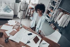 haar ontwerpen zijn perfect. top visie van mooi jong Afrikaanse vrouw glimlachen en op zoek Bij camera terwijl leunend Aan de bureau in werkplaats foto