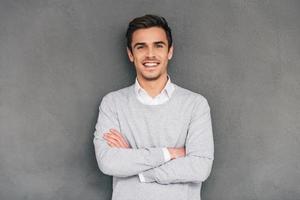 houden lachend. zelfverzekerd jong Mens houden armen gekruiste en op zoek Bij camera met glimlach terwijl staand tegen grijs achtergrond foto