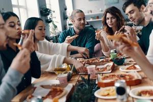 sharing Super goed maaltijd. groep van jong mensen in gewoontjes slijtage aan het eten pizza en glimlachen terwijl hebben een avondeten partij binnenshuis foto