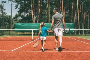 klaar naar Speel. vol lengte achterzijde visie van weinig blond haar- meisje in sport- kleding draag- tennis racket en op zoek Bij haar vader wandelen in de buurt haar door tennis rechtbank foto