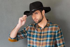 cowboy stijl. knap jong Mens aanpassen zijn cowboy hoed en op zoek Bij camera terwijl staand tegen grijs achtergrond foto