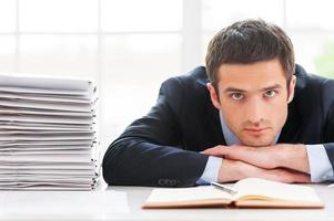 overwerkt leidinggevend. depressief jong Mens in formele kleding op zoek Bij camera en leunend zijn gezicht Bij de tafel met stack van documenten houdende in de buurt hem foto