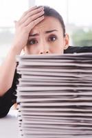 moe van papierwerk. depressief jong vrouw in pak op zoek uit van de stack van documenten houdende Aan de tafel en aanraken haar voorhoofd met hand- foto