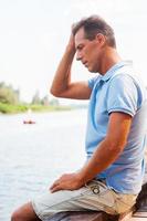 bezorgd en depressief Mens. kant visie van depressief volwassen Mens aanraken hoofd met hand- en op zoek naar beneden terwijl zittend Bij de kade foto