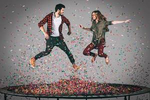 genieten van confetti plezier. in de lucht schot van mooi jong vrolijk paar jumping Aan trampoline samen met confetti allemaal in de omgeving van hen foto