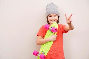 skateboarden is een manier van leven. weinig jongen in hoed Holding kleurrijk skateboard en glimlachen terwijl staand tegen grijs achtergrond foto