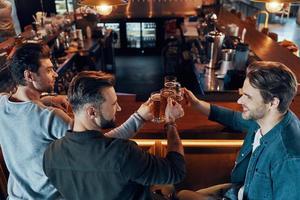 top visie van gelukkig jong mannen in gewoontjes kleding roosteren elk andere met bier en glimlachen terwijl zittend in de kroeg foto