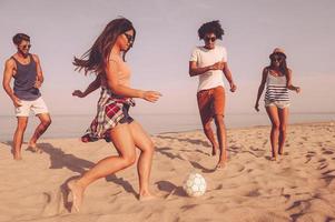 genieten van tijd met het beste vrienden. groep van vrolijk jong mensen spelen met voetbal bal Aan de strand met zee in de achtergrond foto