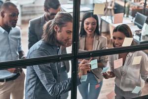 gelukkig bedrijf partner. top visie van jong modern mensen in slim gewoontjes slijtage gebruik makend van Zelfklevend aantekeningen terwijl staand achter de glas muur in de bord kamer foto