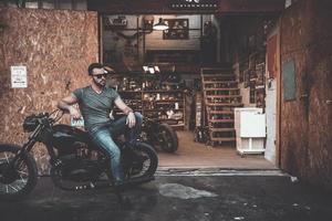 Fietsen zijn niet voor iedereen. knap jong Mens zittend Aan zijn fiets met motorfiets garage in de achtergrond foto