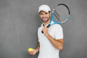 deze spel is meer dan hobby gelukkig jong Mens in sport- kleren draag- tennis racket Aan zijn schouder en glimlachen terwijl staand tegen grijs achtergrond foto