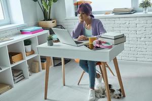 geconcentreerd jong vrouw met Purper haar- werken Aan laptop terwijl zittend Bij haar werken plaats foto