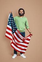 vol lengte van gelukkig jong Afrikaanse Mens gedekt in Amerikaans vlag op zoek Bij camera en glimlachen terwijl staand tegen bruin achtergrond foto