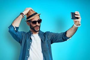 mooi zo op zoek jong Mens glimlachen en nemen selfie gebruik makend van telefoon terwijl staand tegen blauw achtergrond foto