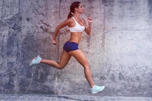 houden Aan rennen. vol lengte kant visie van mooi jong vrouw in sport- kleding rennen met een beton muur in de achtergrond foto