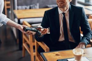 detailopname van knap jong glimlachen Mens in vol pak maken een contactloos betaling terwijl zittend in de restaurant foto