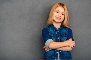 weinig schattig. vrolijk weinig meisje Holding armen gekruiste en op zoek Bij camera met glimlach terwijl staand tegen grijs achtergrond foto