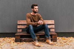 attent knap. knap jong Mens zittend Aan de houten pallet en op zoek weg met grijs muur in de achtergrond en oranje gedaald bladeren Aan de verdieping foto