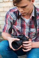 fotograaf onderzoeken camera. knap jong Mens onderzoeken zijn digitaal camera terwijl zittend buitenshuis foto