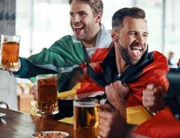 gelukkig jong mannen gedekt in Internationale vlaggen roosteren met bier terwijl aan het kijken sport spel in de kroeg foto