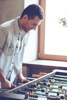 een meer doel. kant visie van vrolijk jong knap Mens spelen tafelvoetbal spel en op zoek opgewonden terwijl staand in voorkant van venster foto