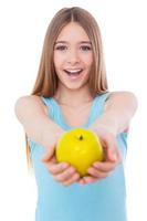 sharing gezond voedsel met jij. vrolijk tiener- meisje uitrekken uit groen appel en glimlachen terwijl staand geïsoleerd Aan wit foto