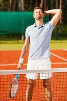 verliezen opnieuw. gefrustreerd jong Mens in sport- kleding aanraken zijn hoofd met hand- en houden ogen Gesloten terwijl Holding tennis racket en staand Aan tennis rechtbank foto