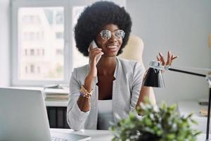 mooi jong Afrikaanse vrouw pratend Aan mobiel telefoon en glimlachen terwijl zittend Bij haar werken plaats in kantoor foto